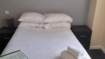A neatly made bed with white linens, a remote control, and rolled towels on the bed.
