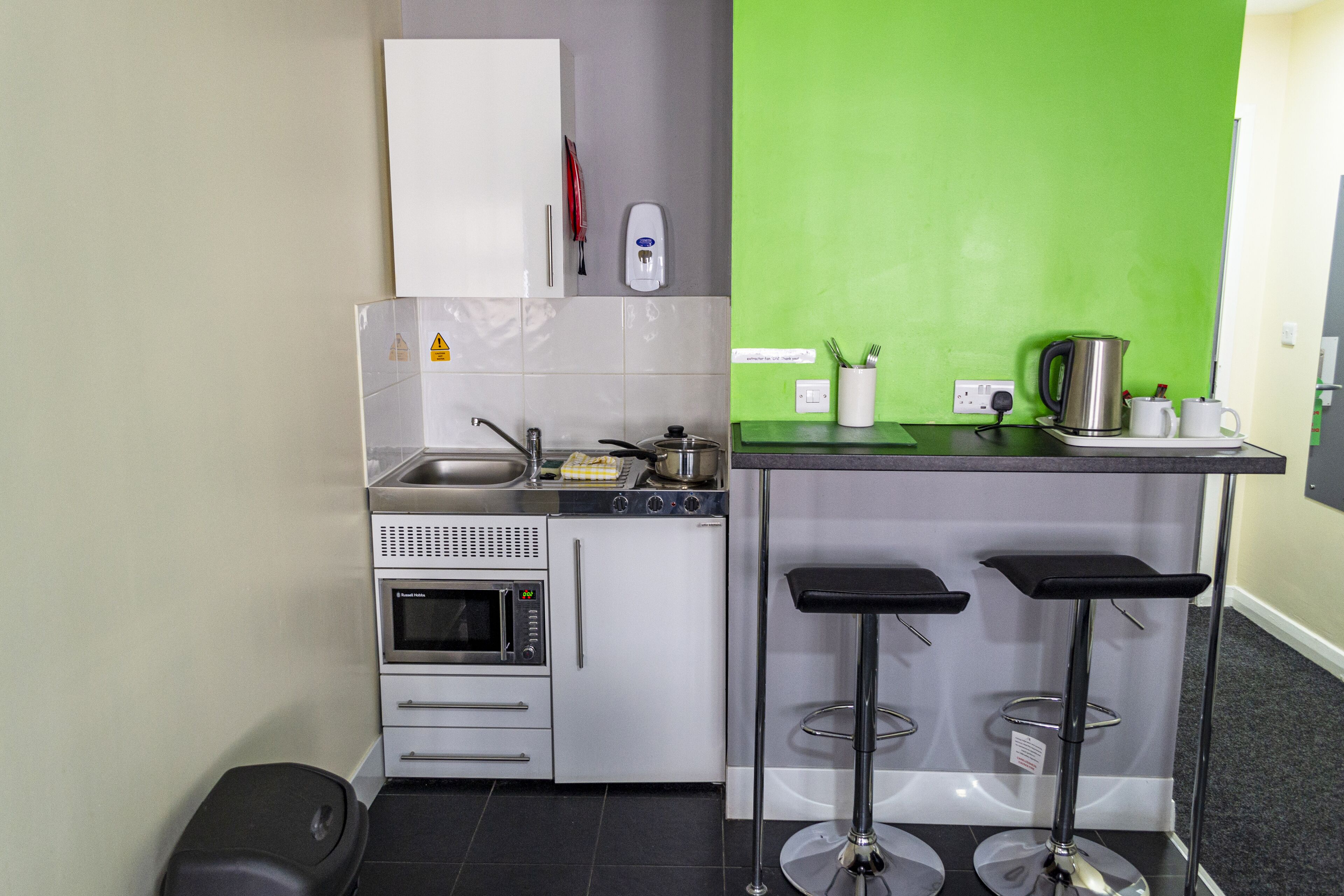 Une cuisine compacte équipée d’un évier, d’une cuisinière, d’un four et d’un mur d’accent vert. On y trouve deux tabourets de bar et un petit comptoir avec une bouilloire et des tasses.