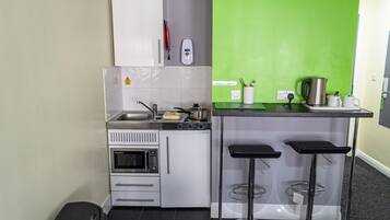 Une cuisine compacte équipée d’un évier, d’une cuisinière, d’un four et d’un mur d’accent vert. On y trouve deux tabourets de bar et un petit comptoir avec une bouilloire et des tasses.