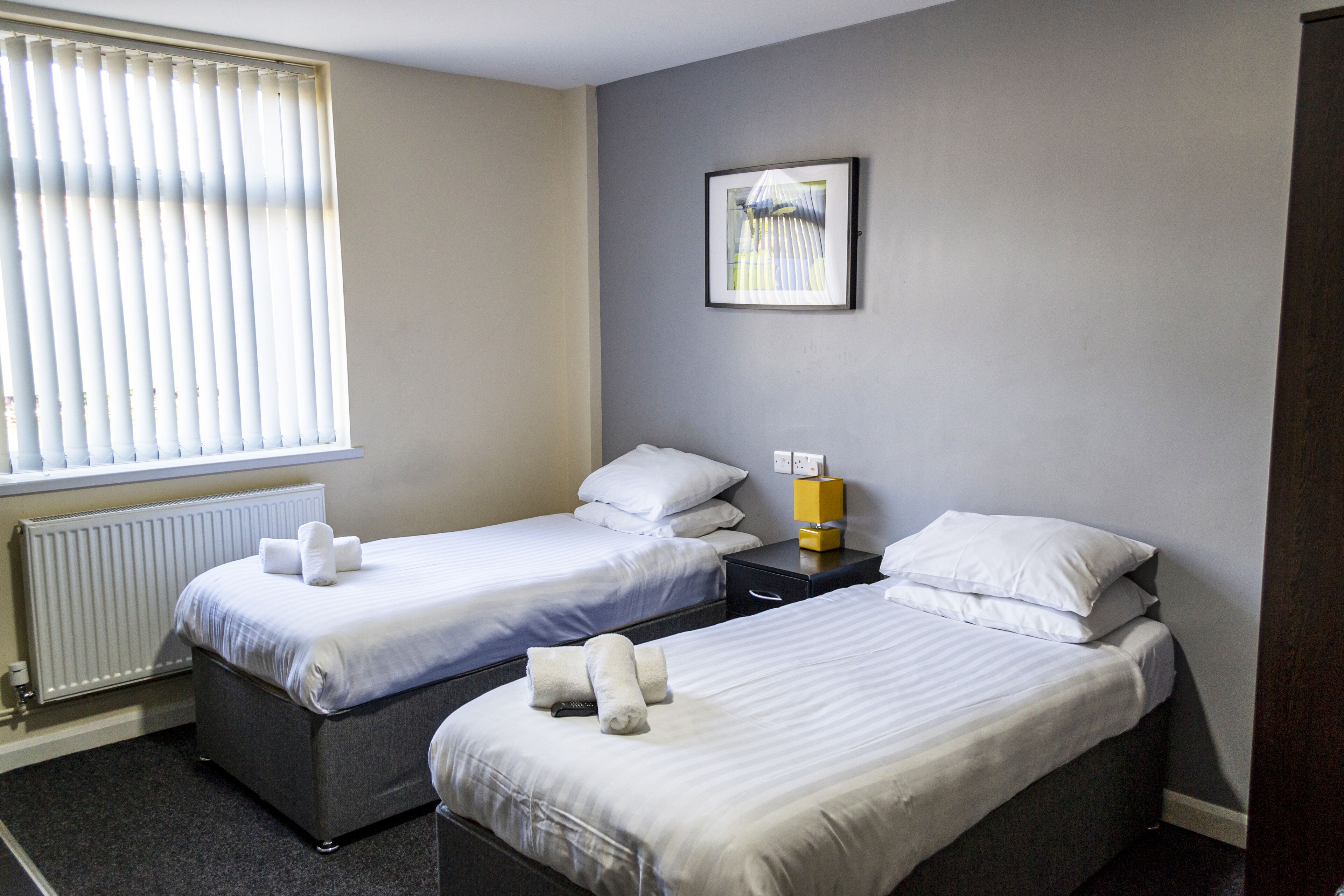 Ein Hotelzimmer mit zwei Betten, einem Heizkörper, einem gerahmten Bild an der Wand und einem Fenster mit Jalousien.