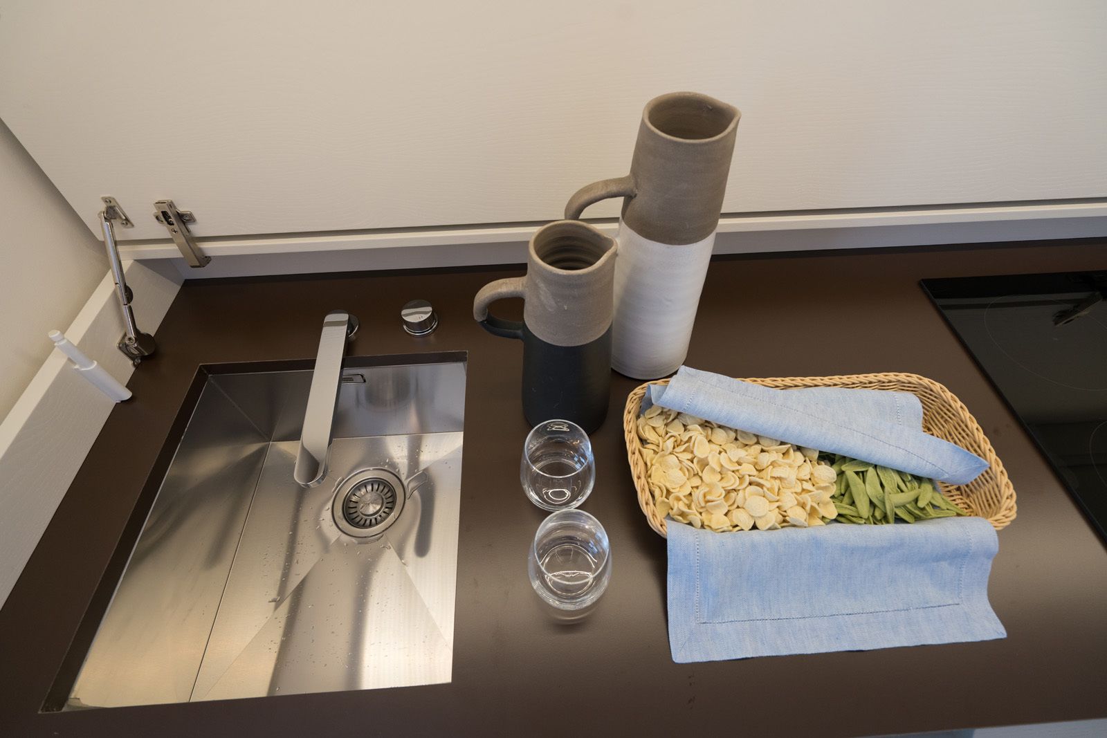 A kitchen counter with a stainless steel sink, a knife, a mortar and pestle, a mortar, a mortar with a lid, and a basket of pasta.