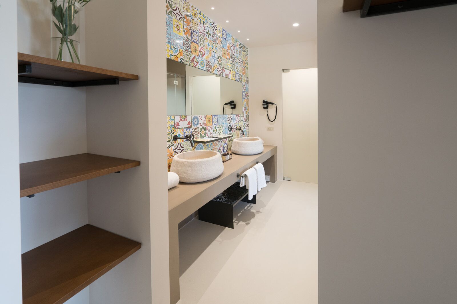 A modern bathroom with two sinks, a mirror, and a shower. There are wooden shelves and a plant on the left.