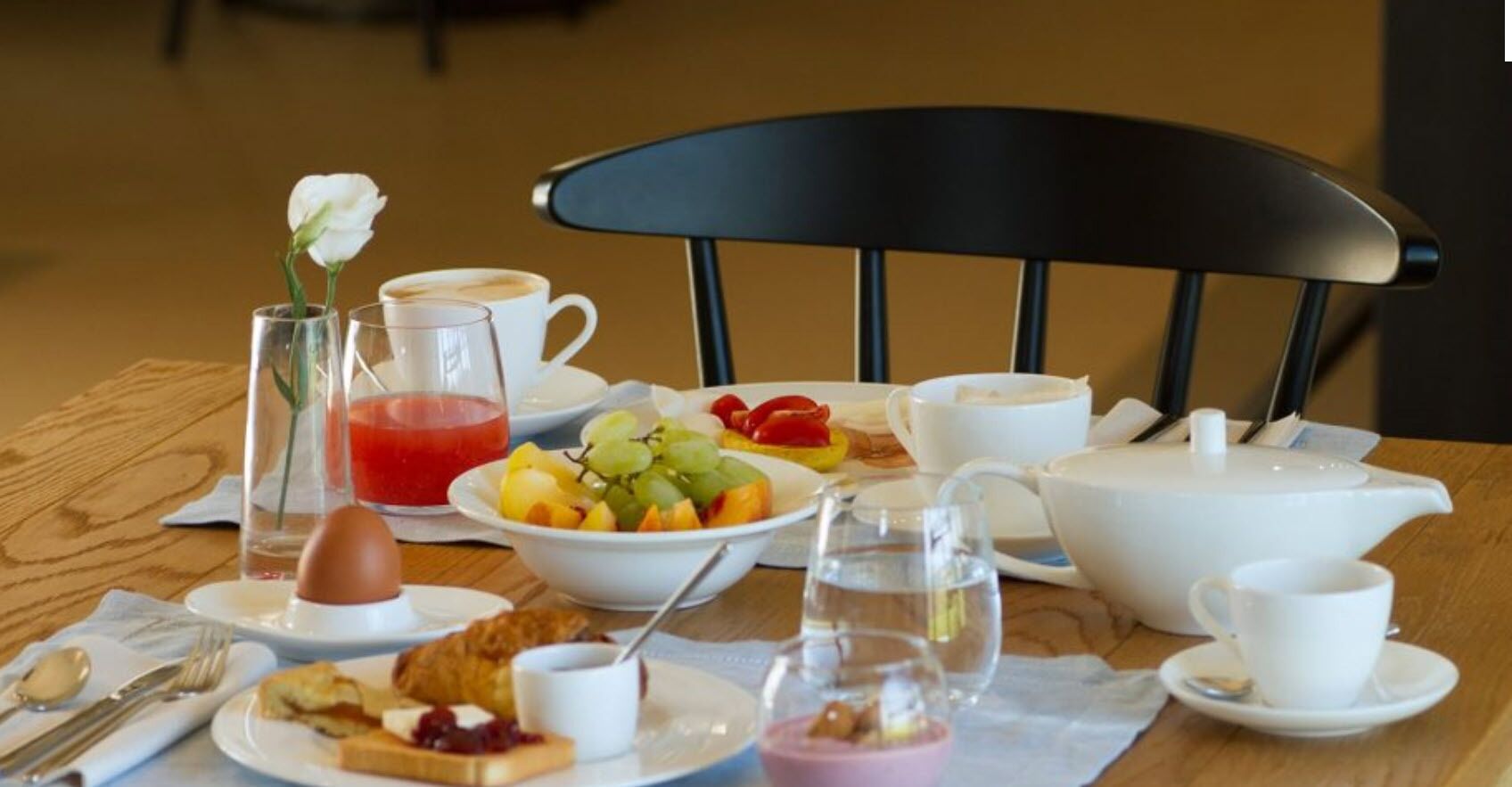 A breakfast table with a variety of food items, including fruits, pastries, and beverages, arranged on a wooden table with a black chair in the background.