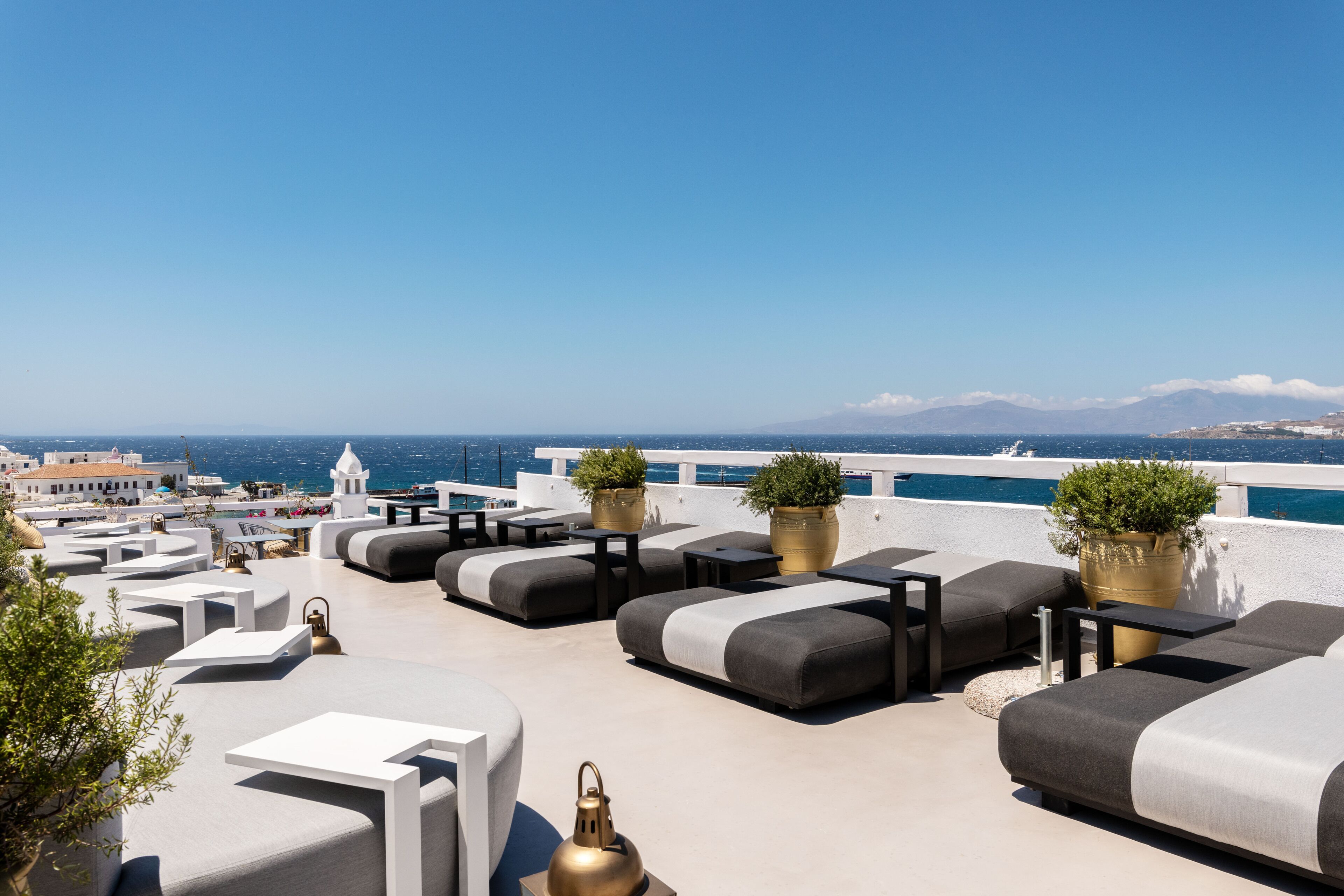 Eine Dachterrasse mit Sitzmöglichkeiten im Freien, Blumentöpfen und Blick auf das Meer und ferne Berge.