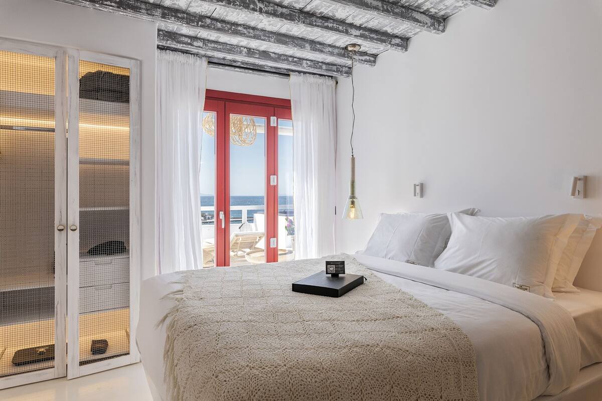 Ein Schlafzimmer mit Bett, Nachttisch, einer roten Tür und Blick aufs Meer.