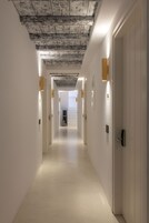 Un pasillo largo y estrecho con paredes blancas y techo de madera. Hay puertas a ambos lados, con pequeñas luminarias rectangulares sobre ellas.