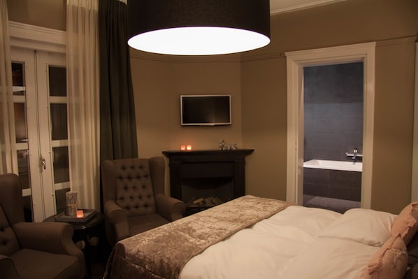 Habitación de hotel con cama, una silla, televisor, chimenea y baño visible a través de una puerta abierta.