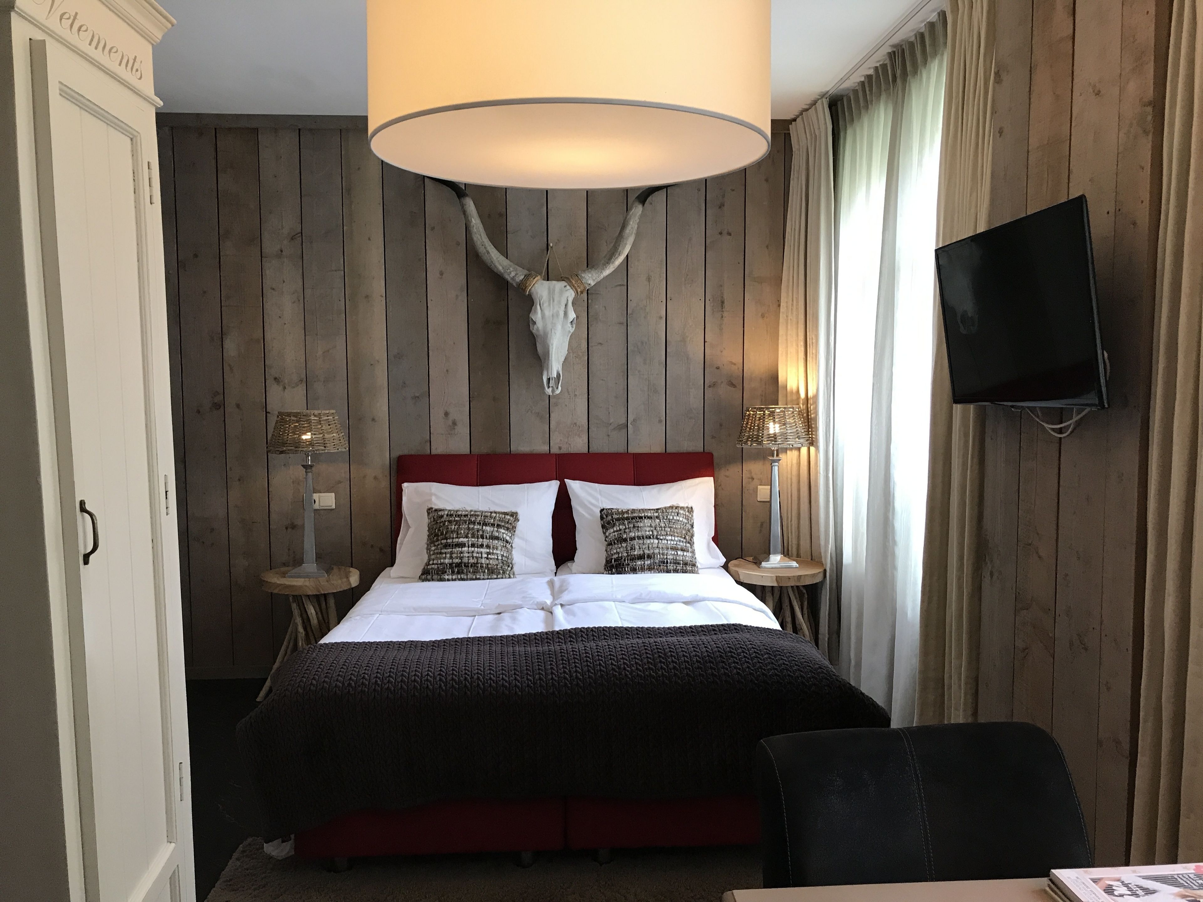 A hotel room with a wooden panel wall, a mounted deer skull, a red upholstered bed, two bedside lamps, a television, and a desk with a chair.