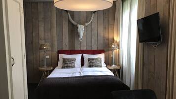A hotel room with a wooden panel wall, a mounted deer skull, a red upholstered bed, two bedside lamps, a television, and a desk with a chair.