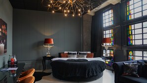 A modern bedroom with a round black bed, two bedside tables with lamps, a black sofa, and large stained glass windows. - Hotel Huis van Bewaring (Almelo)