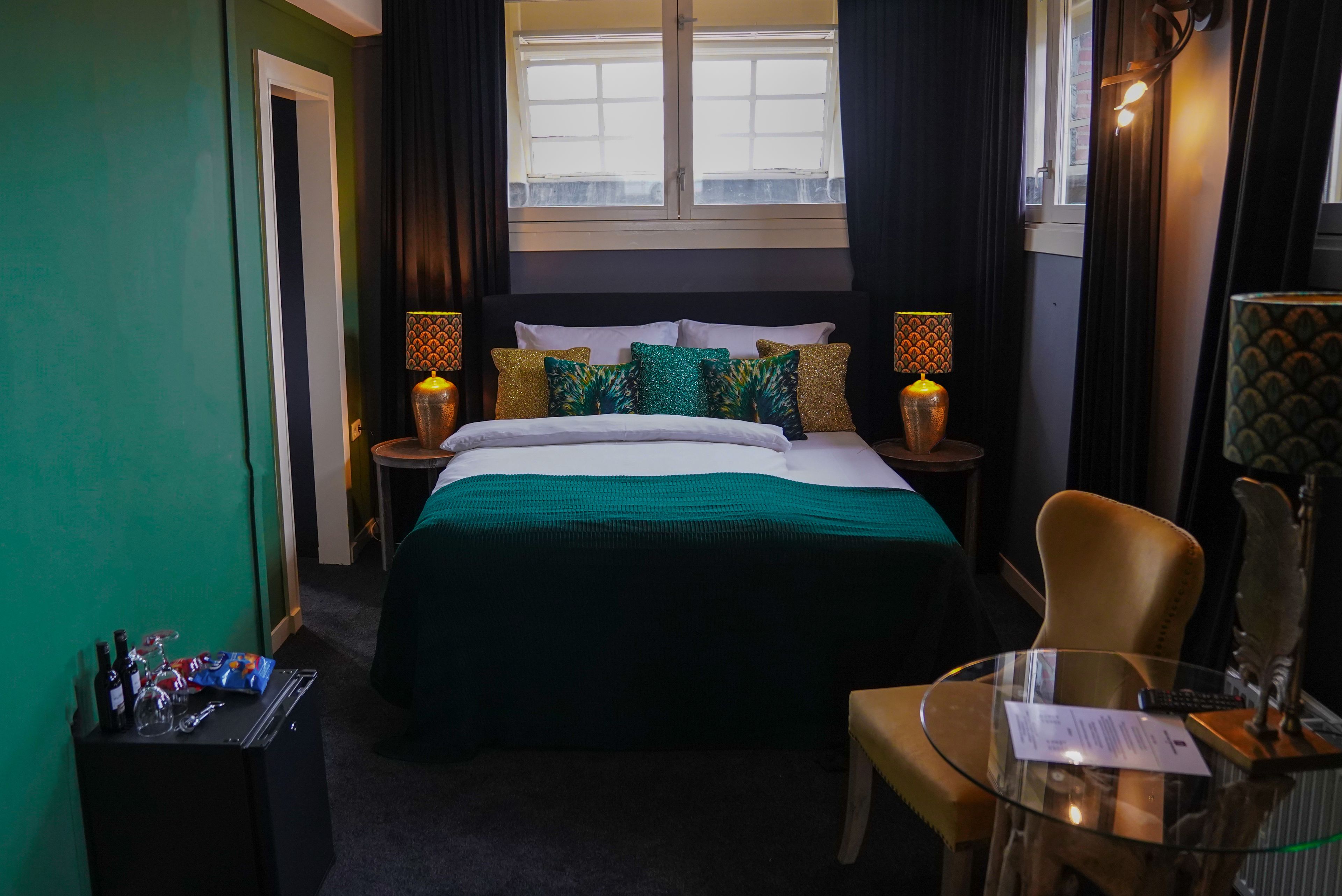 A hotel room with a bed, a glass table, a chair, a nightstand with a lamp, and a window with curtains.