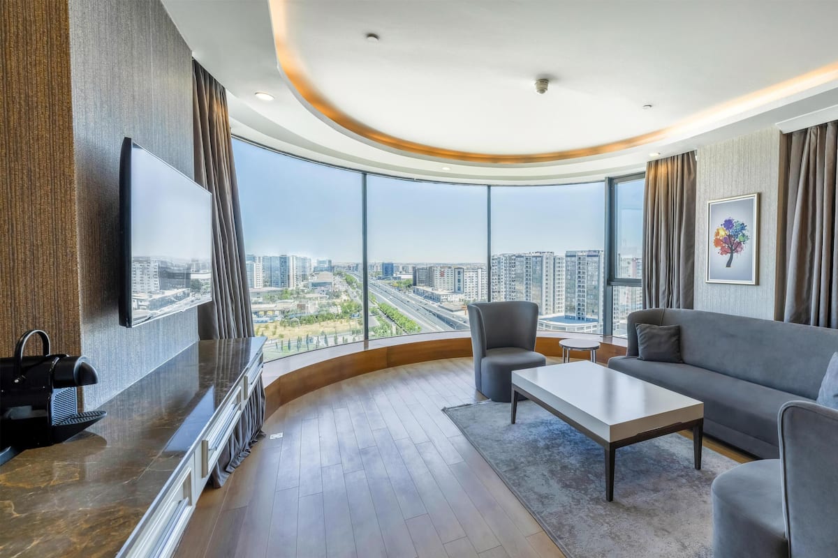 Una camera d'albergo moderna con soffitto curvo, un'ampia finestra con vista sulla città, un divano grigio, un tavolino e una sedia.
