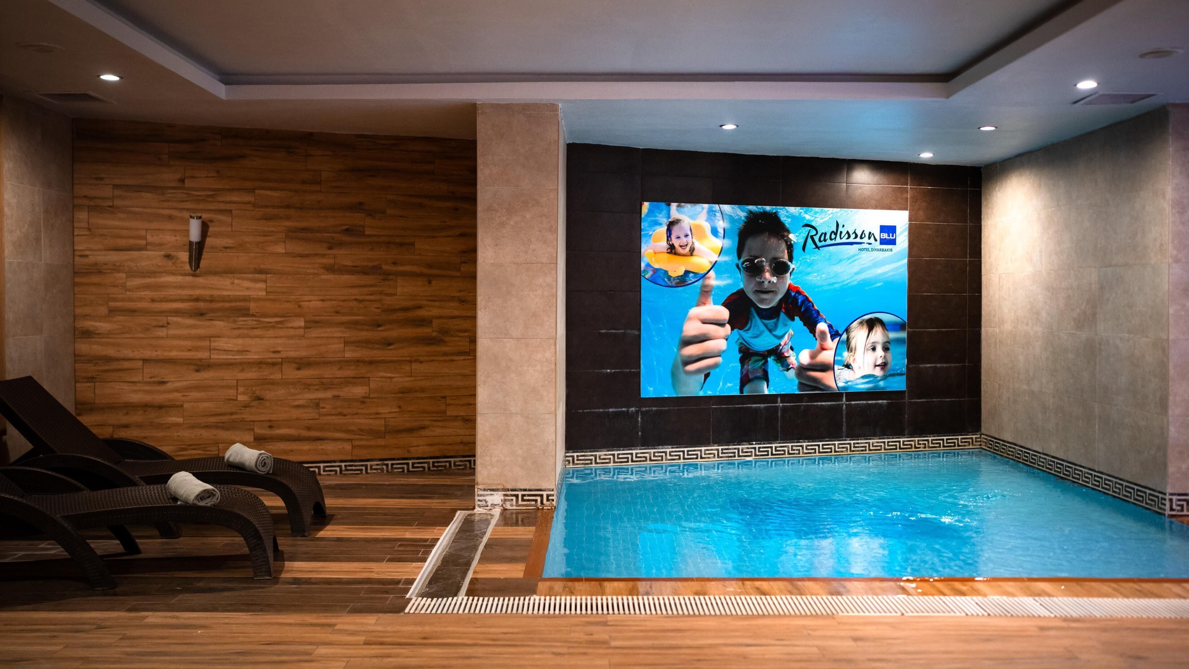 Ein Poolbereich mit Holzboden, Liegestühlen und einem großen Bildschirm, der eine Werbung zeigt.
