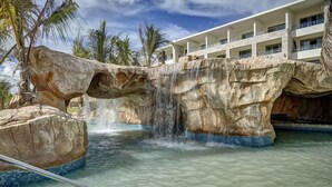 A pool area with a waterfall feature, large rock formations, and a modern building in the background. - Royalton Bavaro (Punta Cana)