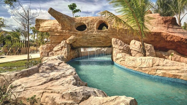 A rock bridge over a pool with clear water, surrounded by palm trees and rocky terrain. - Royalton Bavaro (Punta Cana)
