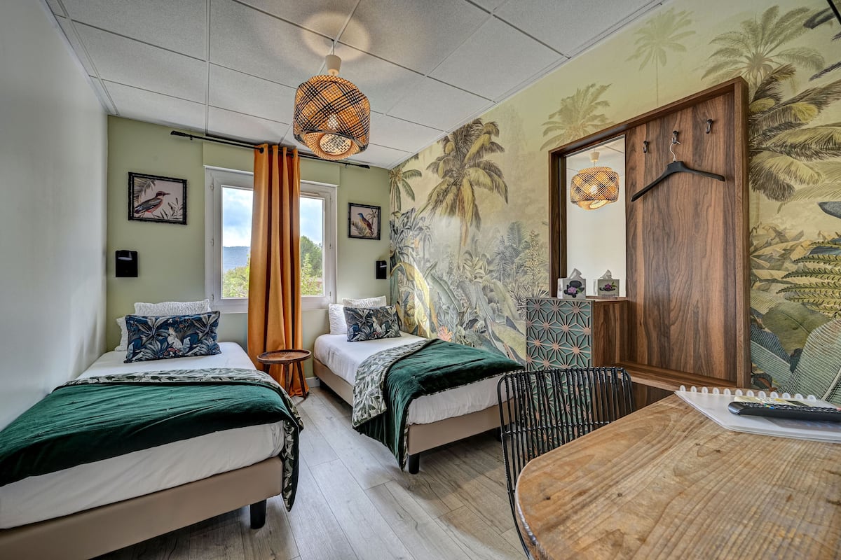 A room with two beds, a wooden table, a hanging light fixture, a door with a wooden panel, and a mural on the wall.