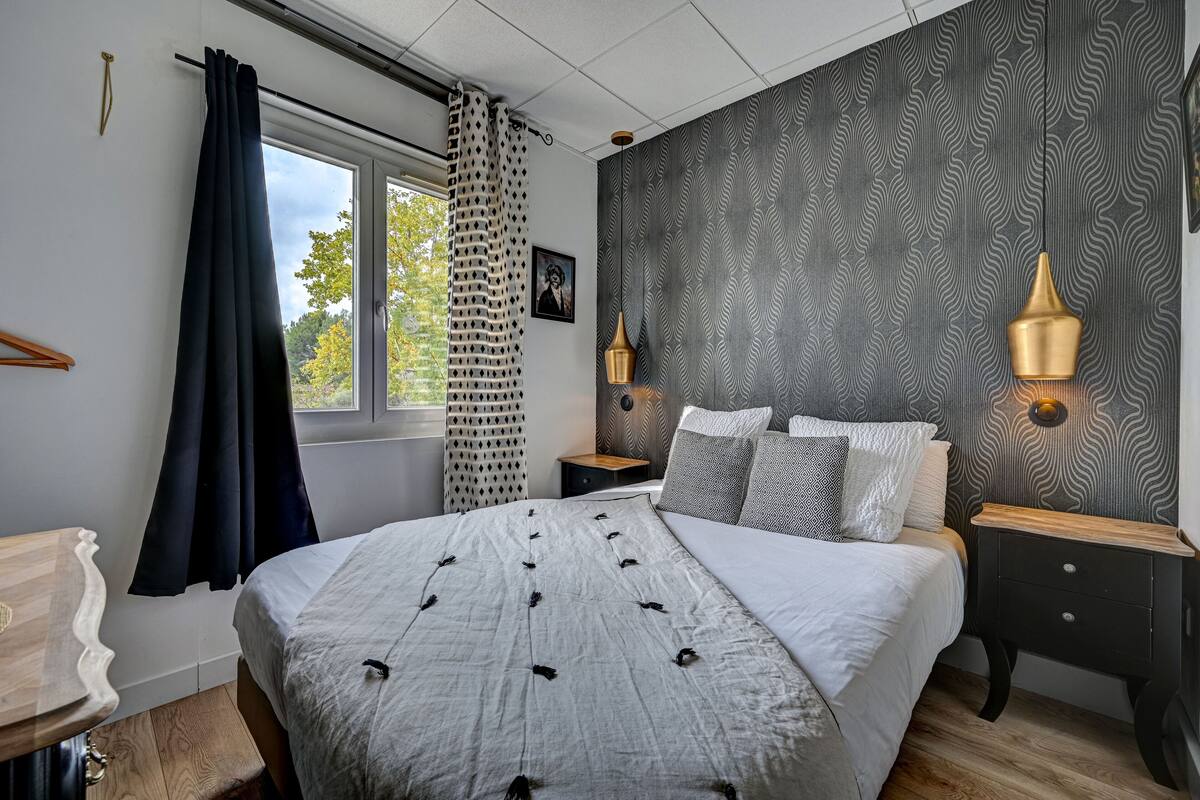 A bedroom with a large bed, two bedside tables with lamps, a window with curtains, and a patterned wall.