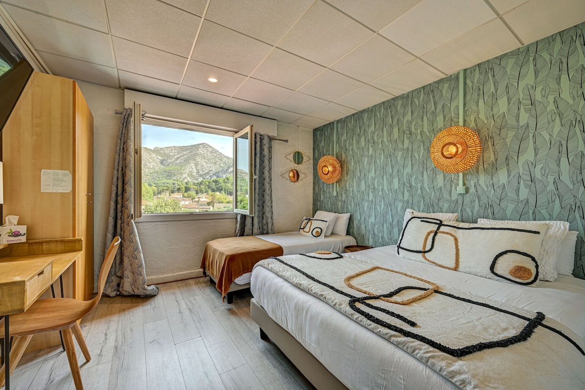 A bedroom with a desk, two beds, a view of mountains, and decorative wall art.