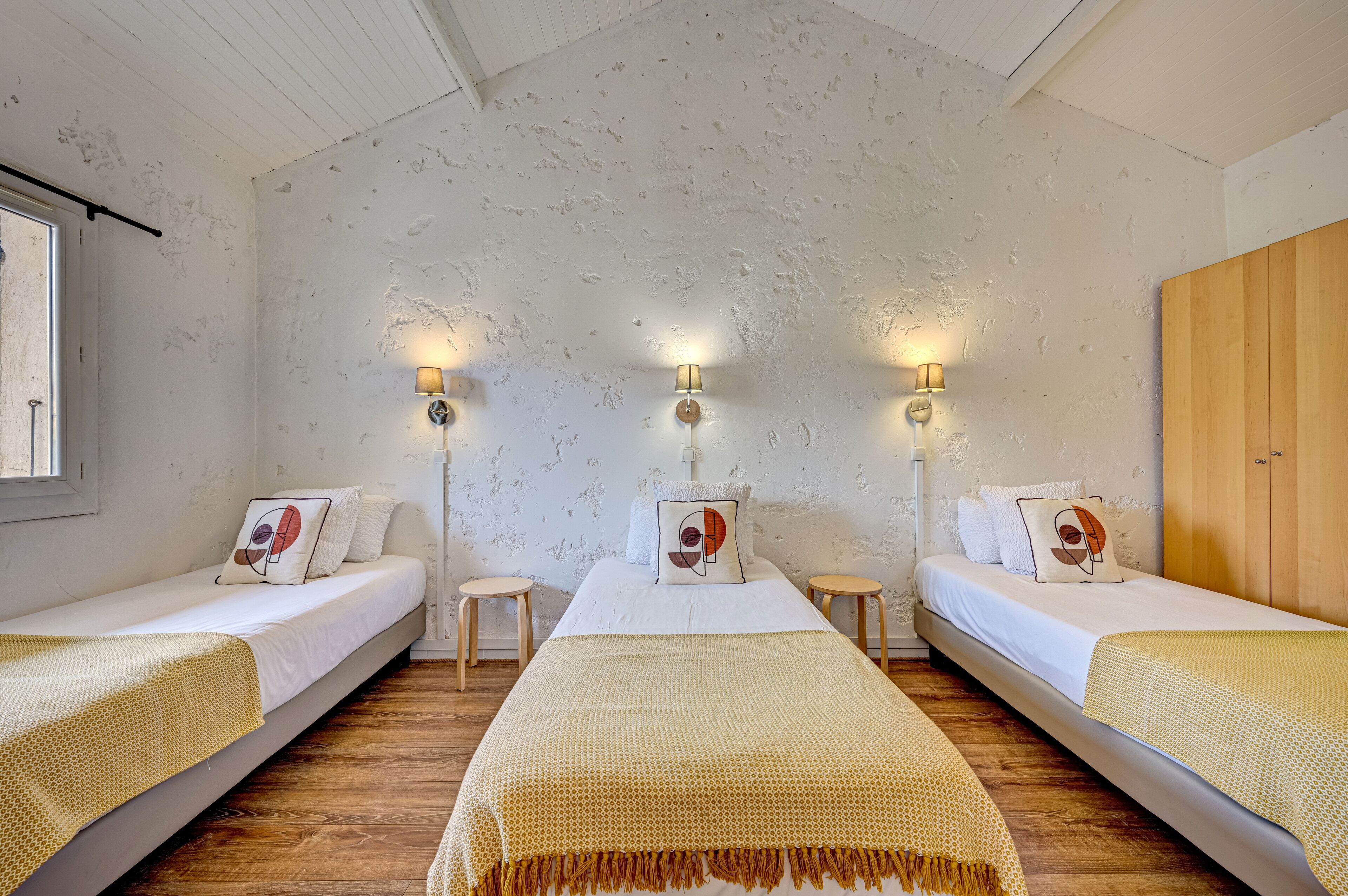 Una habitación con tres camas individuales, cada una con mesita de noche y lámpara. Las paredes tienen una textura decorativa y hay un armario de madera a la derecha.