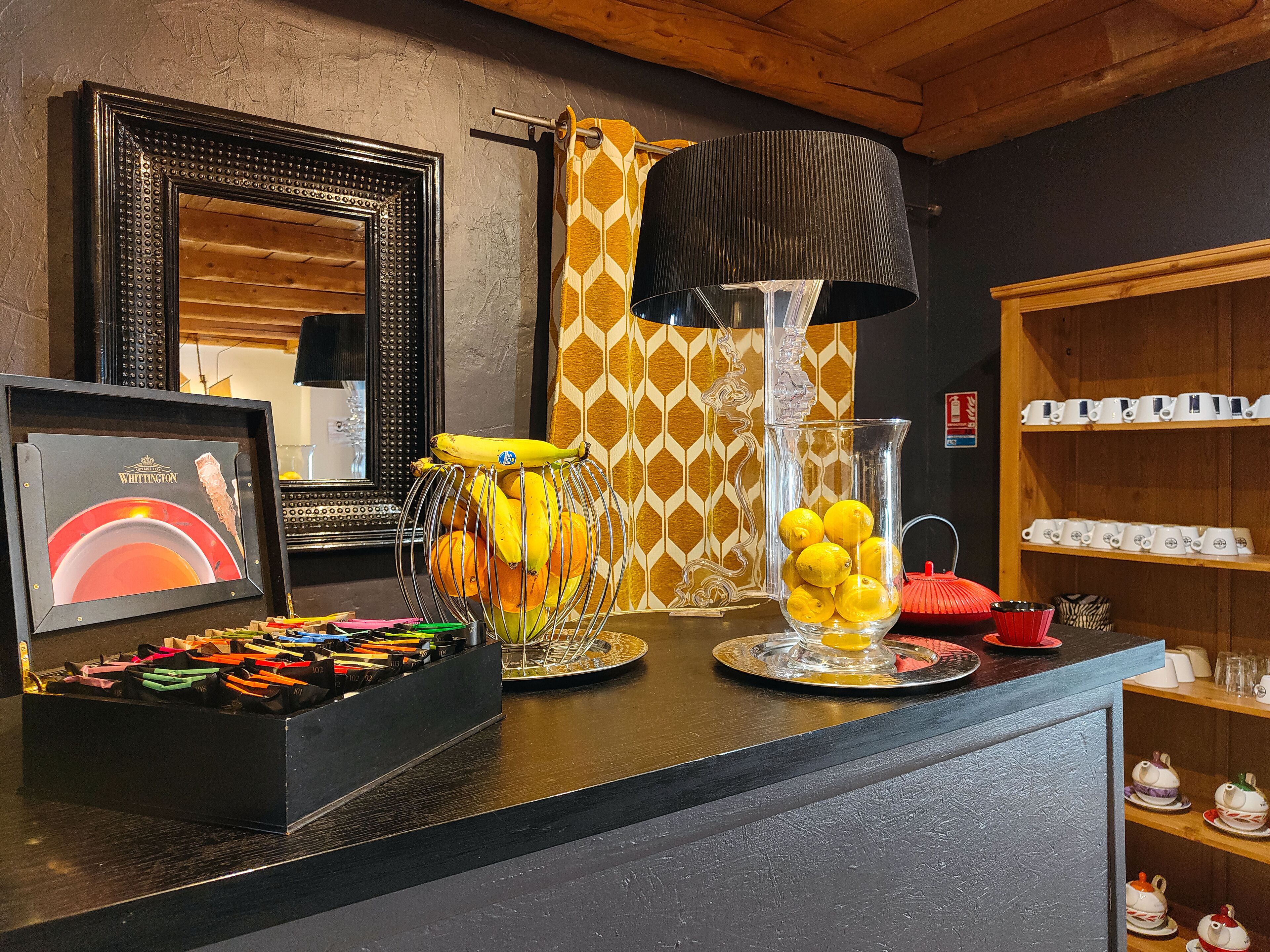 A bar area with a black countertop, a framed picture, a metal fruit basket, a glass pitcher with lemons, and shelves with white cups.