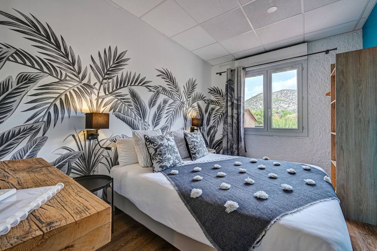 A bedroom with a bed, a wooden nightstand, a wall-mounted lamp, and a window with a view of greenery.