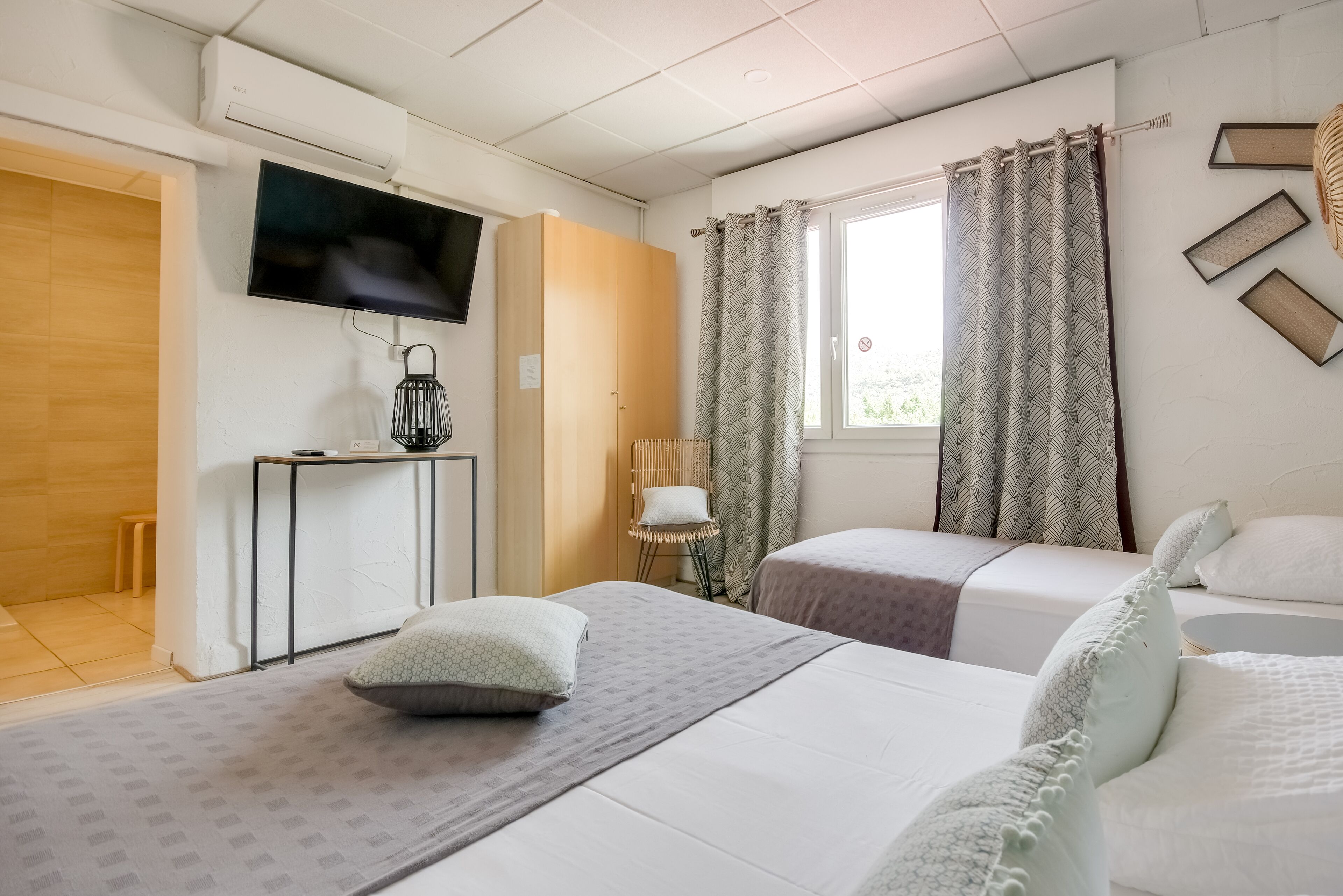 A hotel room with two beds, a TV mounted on the wall, a small table with a lamp, and a window with curtains.