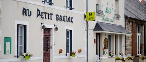 A building with a sign reading "Au Petit Breuil" and another sign for "Hotel Restaurant". There are flower pots on the sidewalk and a chair outside the building.