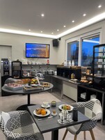 Espaço de café da manhã moderno com buffet, mesas e cadeiras, e uma TV exibindo uma vista da cidade.