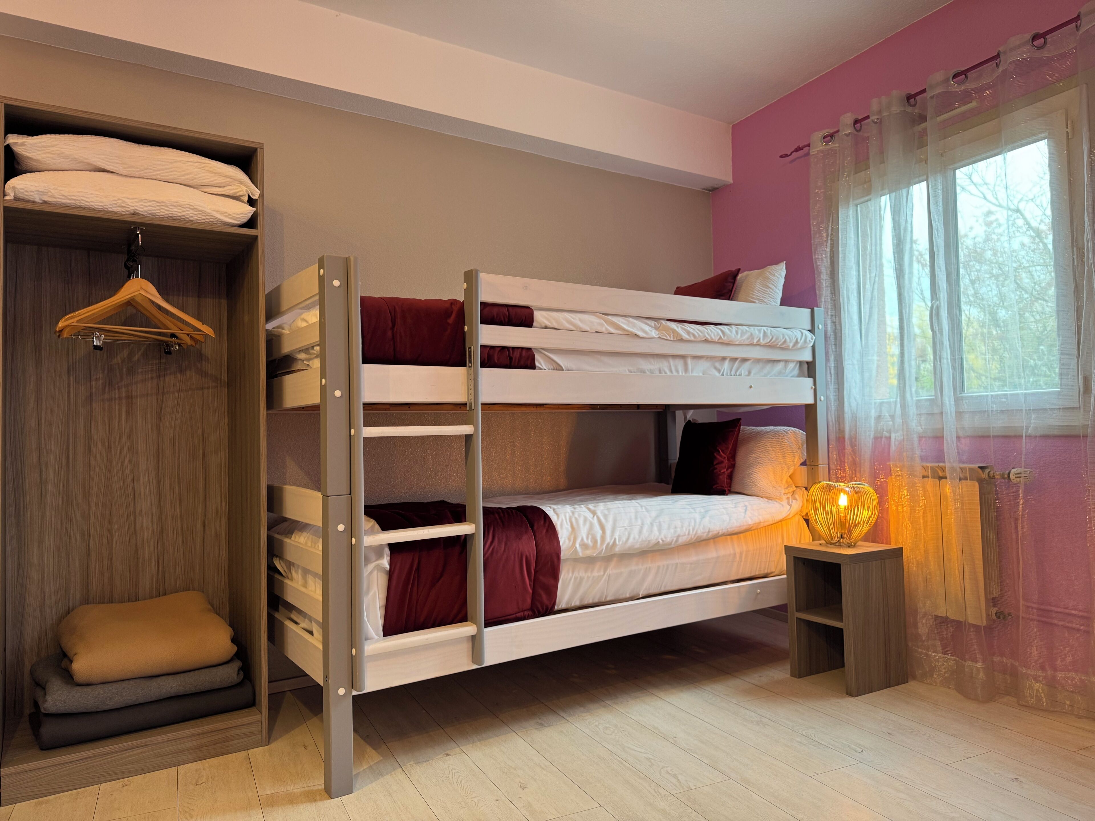 Een slaapkamer met een stapelbed, een houten garderobe, een klein nachtkastje met lamp en een raam met gordijnen.