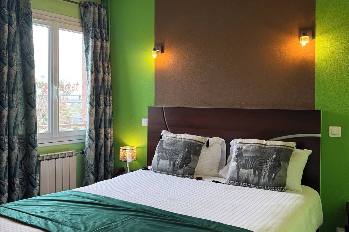 A hotel room with a large bed, green walls, zebra-themed pillows, a nightstand with a lamp, and a window with curtains.