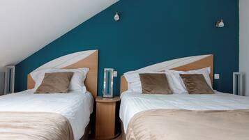A bedroom with two beds, each with a wooden headboard, white bedding, and brown pillows. There are bedside tables with lamps and a mirror. The room has a sloped ceiling and teal walls.