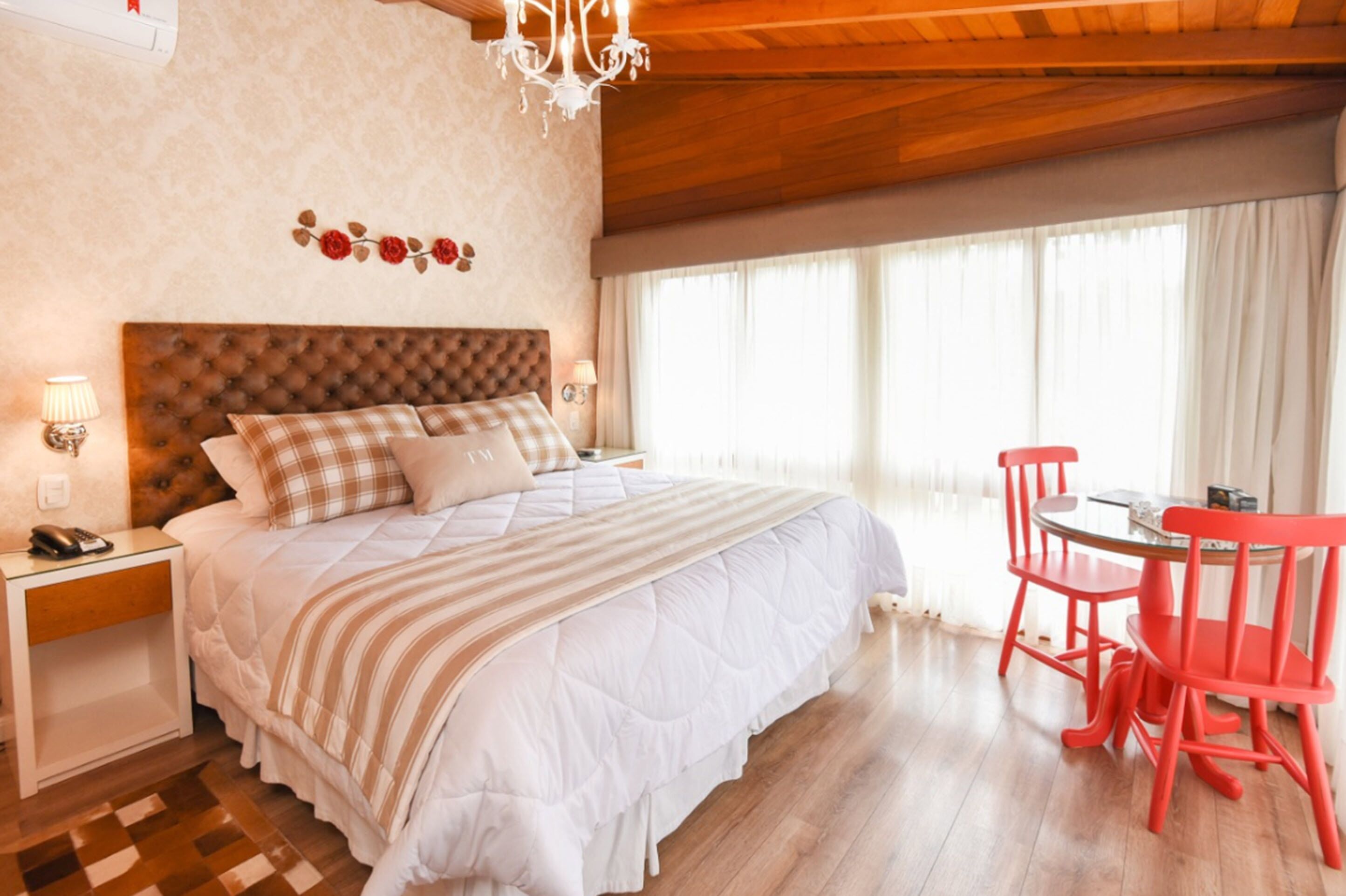 A bedroom with a large bed, a wooden ceiling, a chandelier, a small table with chairs, and a window with curtains.