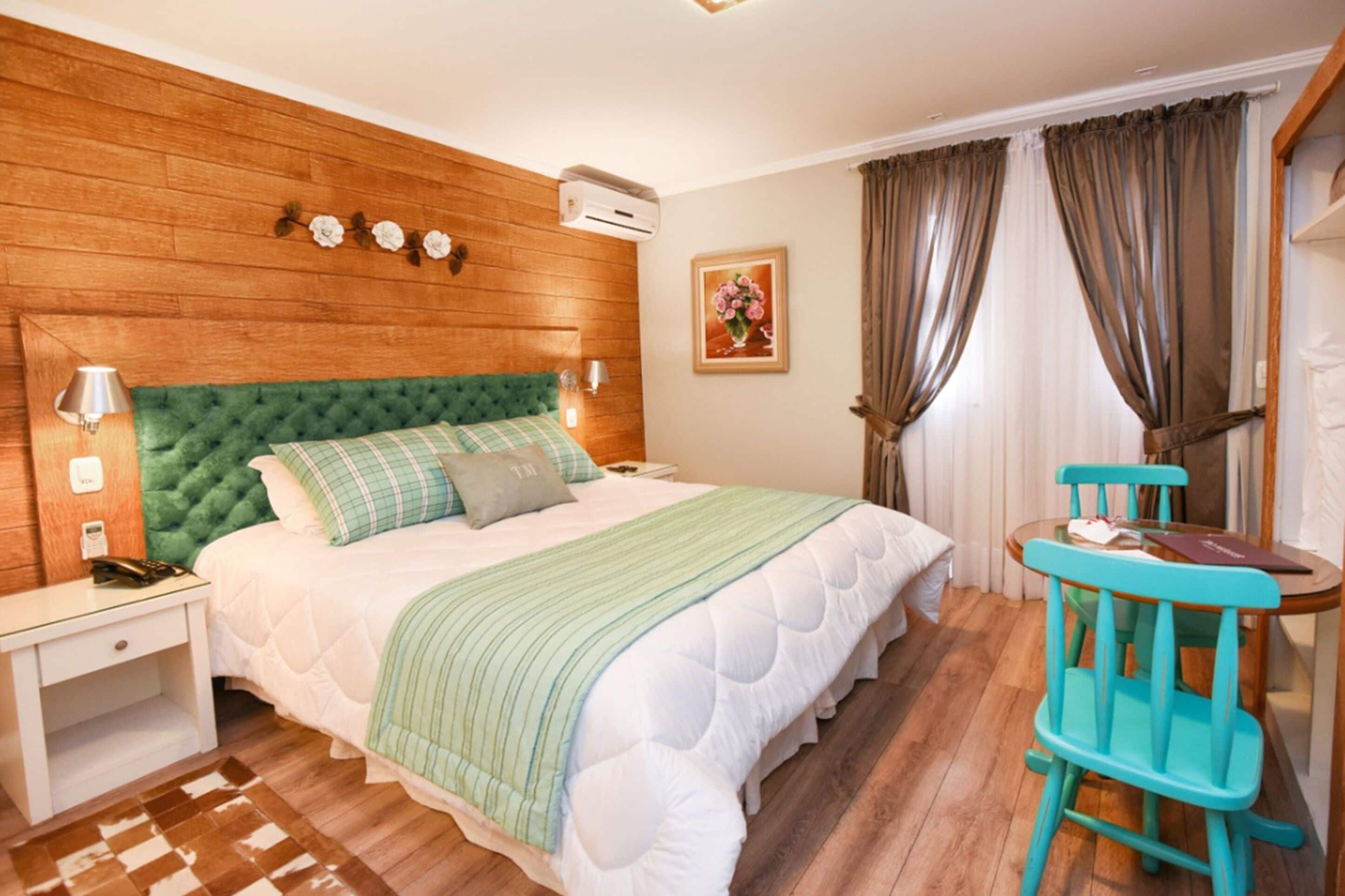 Ein Schlafzimmer mit einem grünen, gesteppten Kopfteil, einem Bett mit weißer Bettwäsche, einer grün-weißen Streifenauflage und einem Nachttisch aus Holz mit einer Lampe, sowie einem türkisfarbenen Sessel.