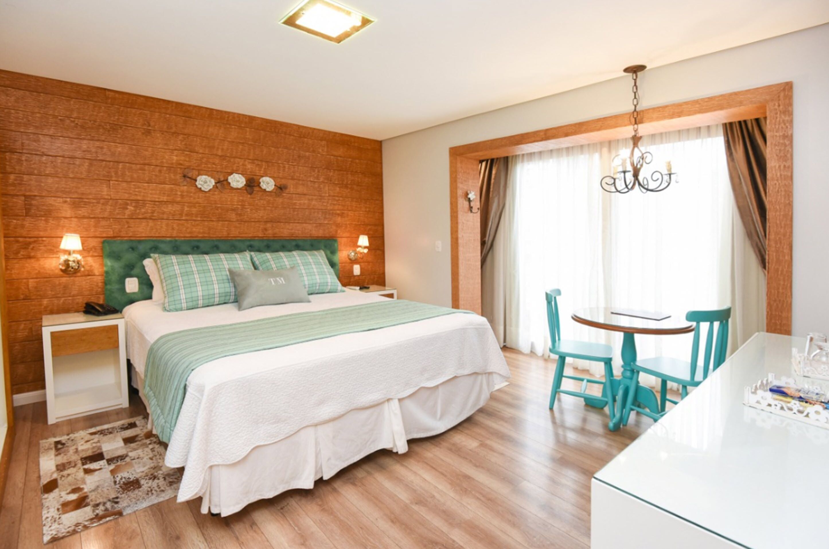 Un dormitorio con cabecera de madera, cama con colcha verde y blanca, dos lámparas de noche, una pequeña mesa con sillas y una araña.