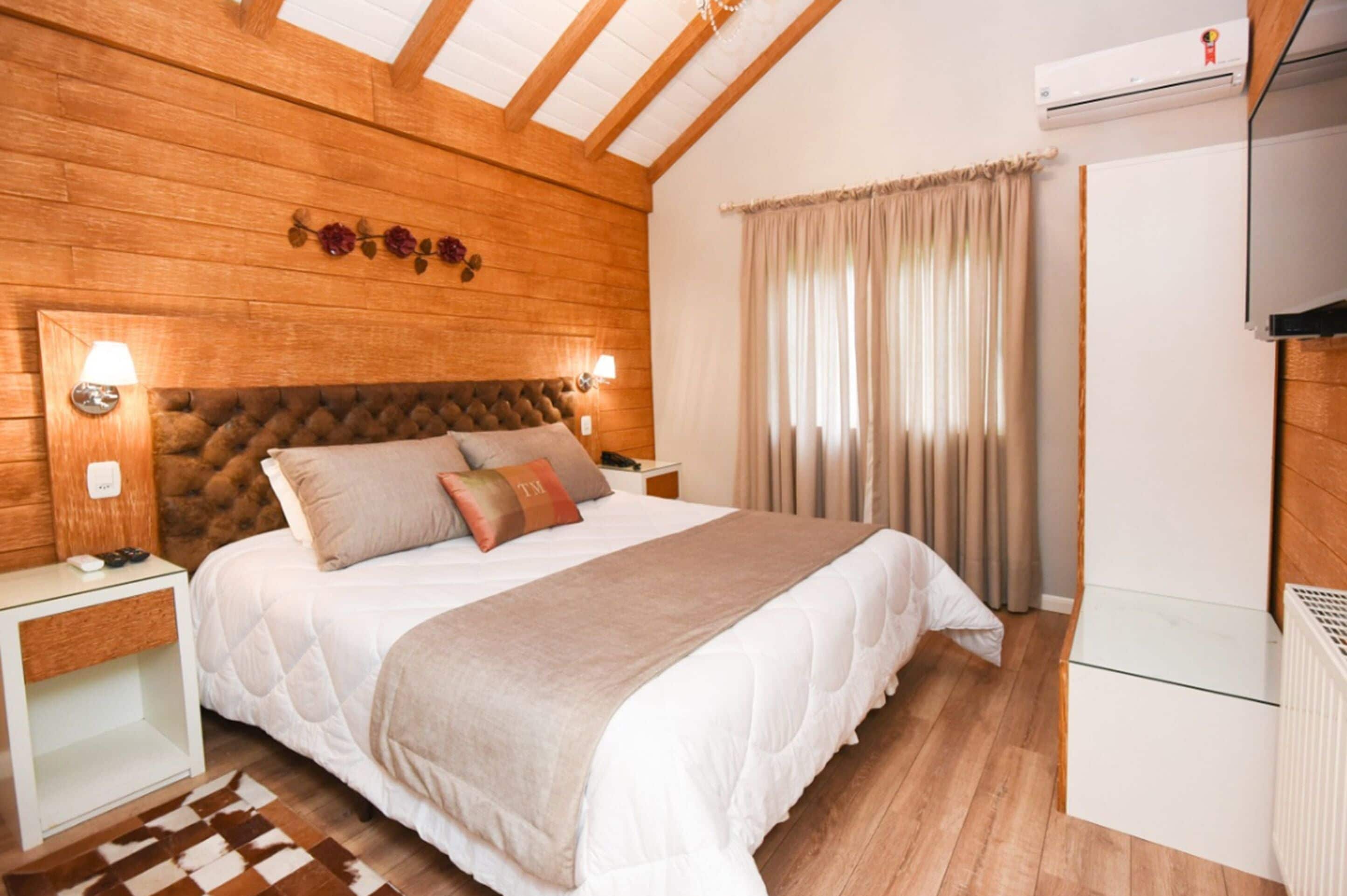 A bedroom with a wooden ceiling, a bed with a white duvet, two bedside lamps, and a window with curtains.