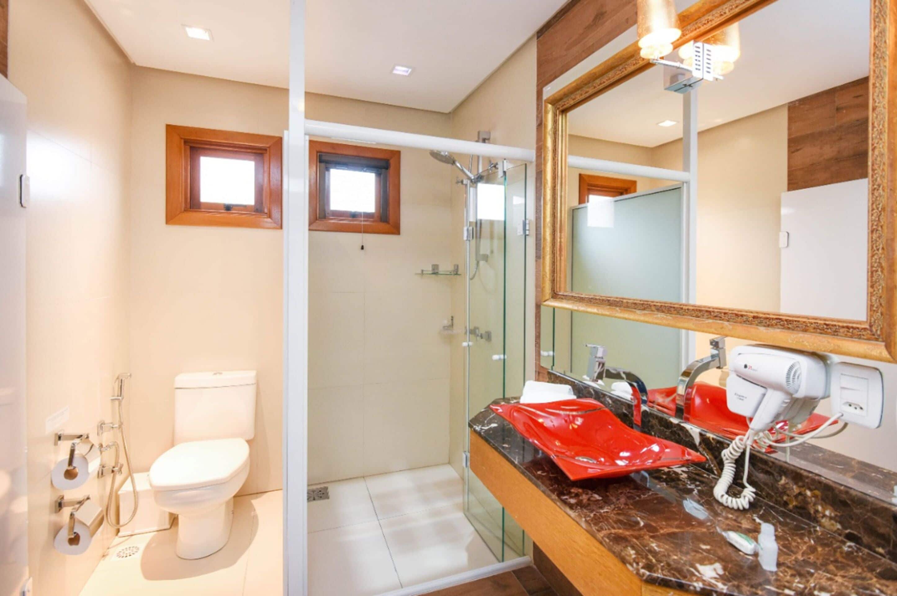 A bathroom with a glass shower enclosure, a white toilet, a vanity with a red sink, and a large mirror.