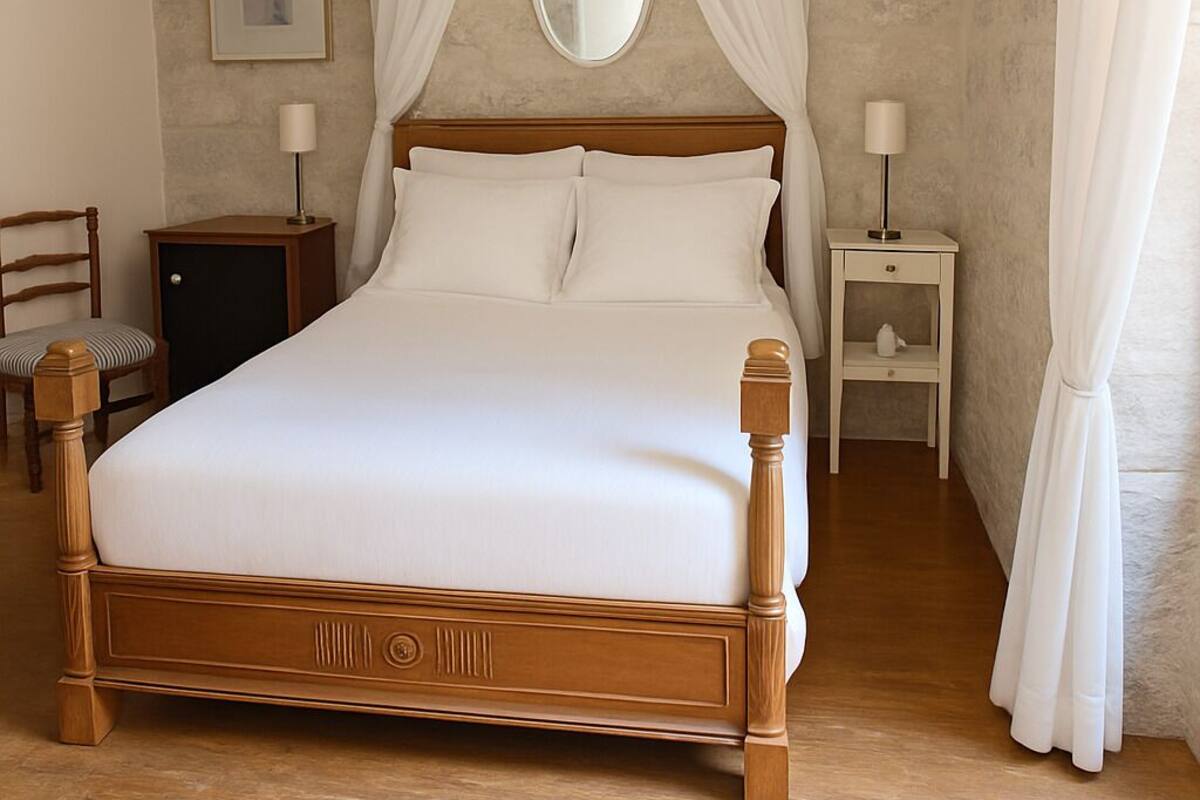 A neatly made bed with a wooden frame and white bedding, a bedside table with a lamp, a chair, and a framed picture on the wall.