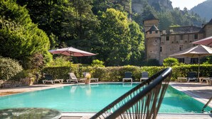 Un área de piscina con sillones de descanso y sombrillas, rodeada de exuberante vegetación y un edificio con aspecto de castillo al fondo.