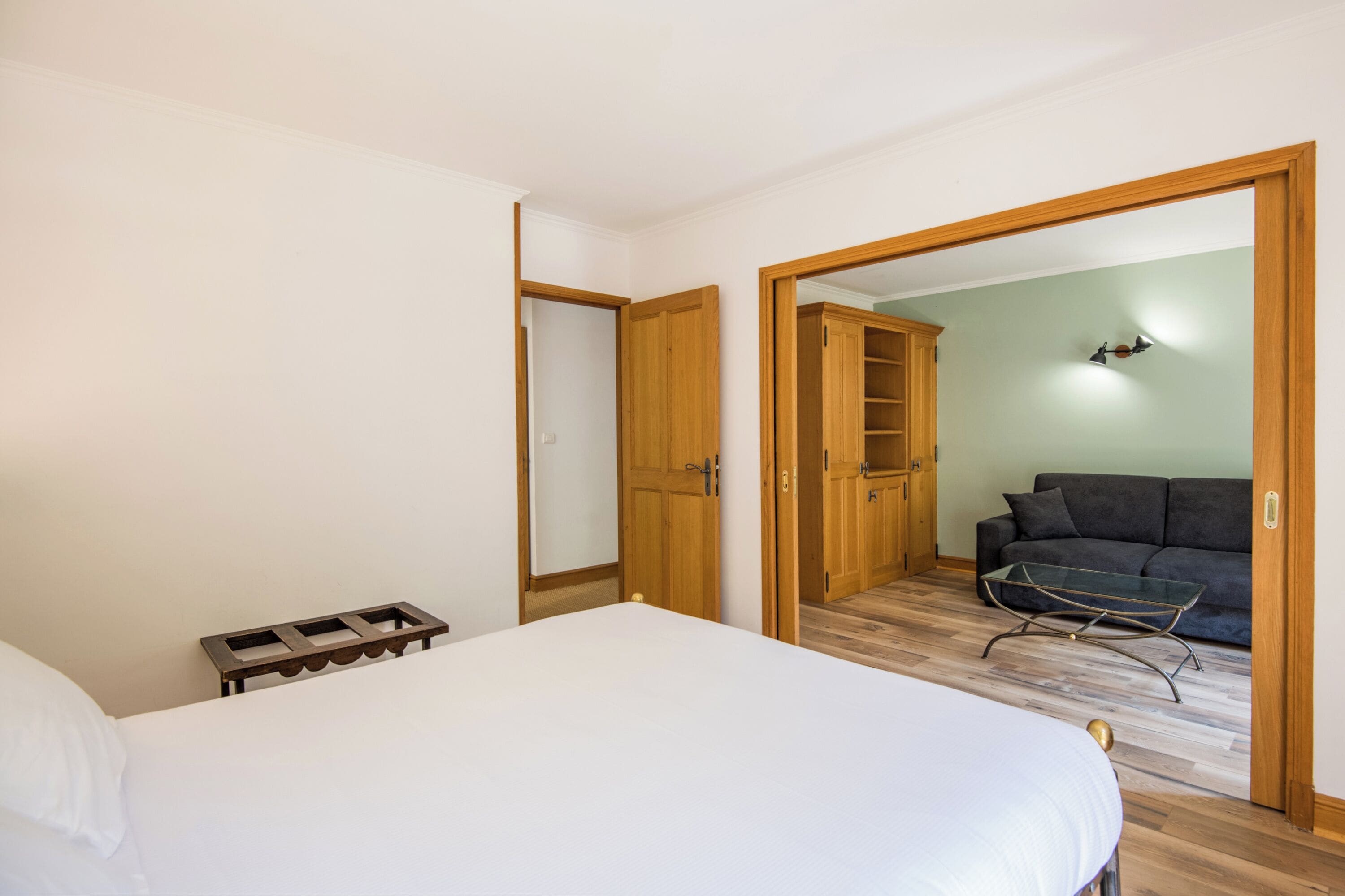 A bedroom with a bed, a bedside table, a wardrobe, and a sofa in the adjacent room.