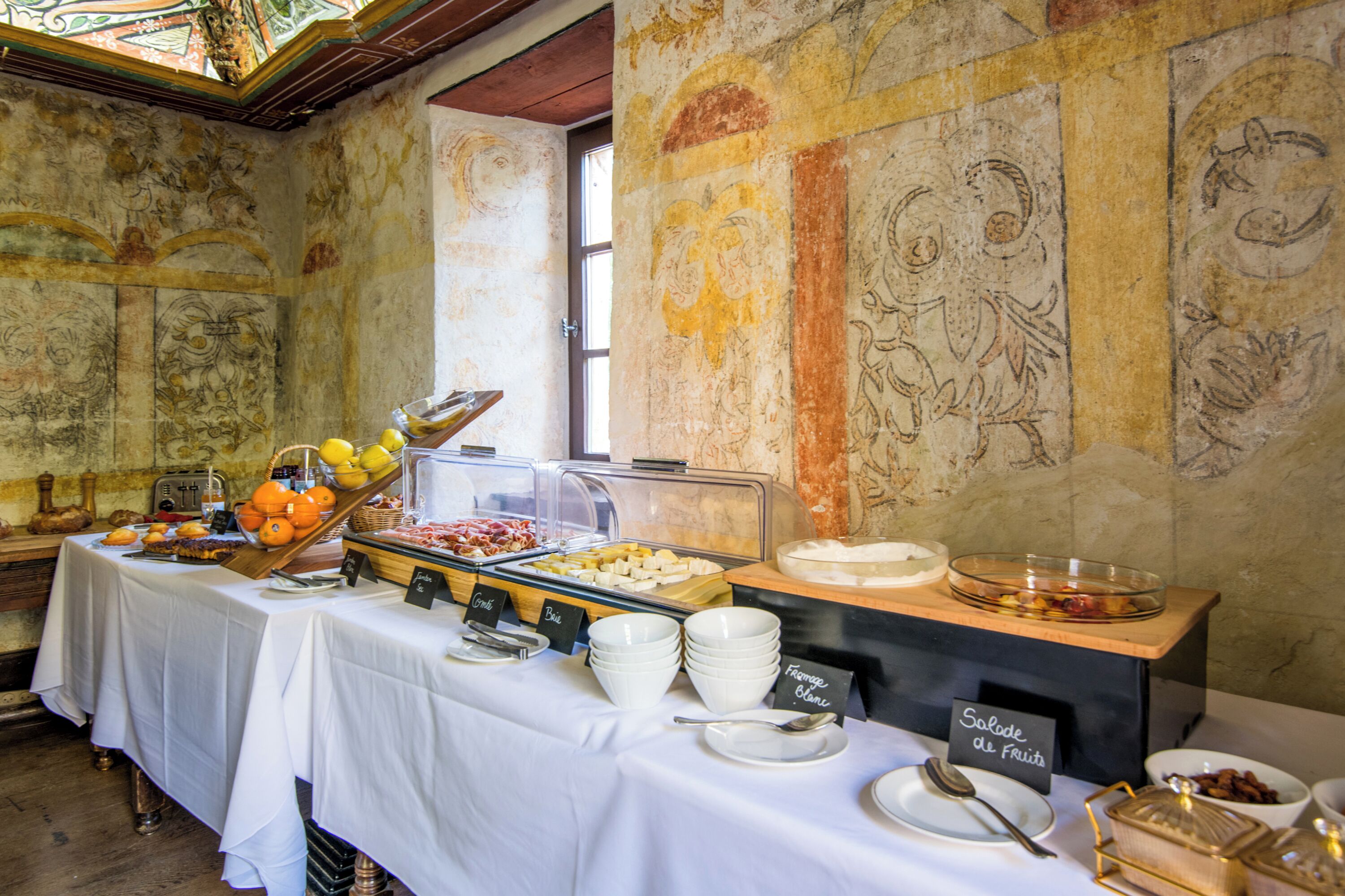 A buffet setup with various food items and labeled containers, set against a backdrop of ornate wall murals.