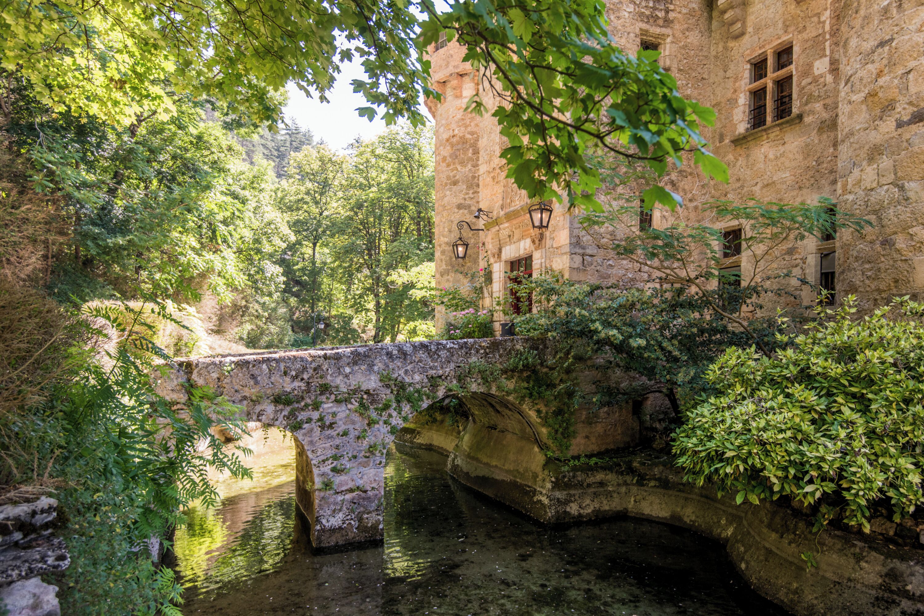 Un puente de piedra sobre un cuerpo de agua, rodeado de abundante vegetación y un edificio histórico con ventanas arqueadas.