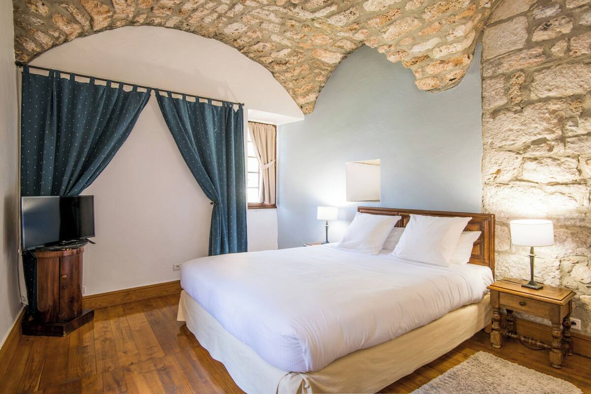 A bedroom with a stone wall, a wooden bed, a nightstand with a lamp, and a TV on a stand.