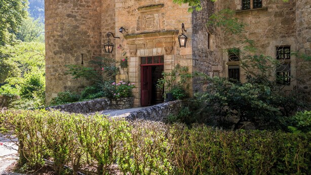 Un edificio de piedra con una puerta roja, rodeado de exuberante vegetación y un muro de piedra.