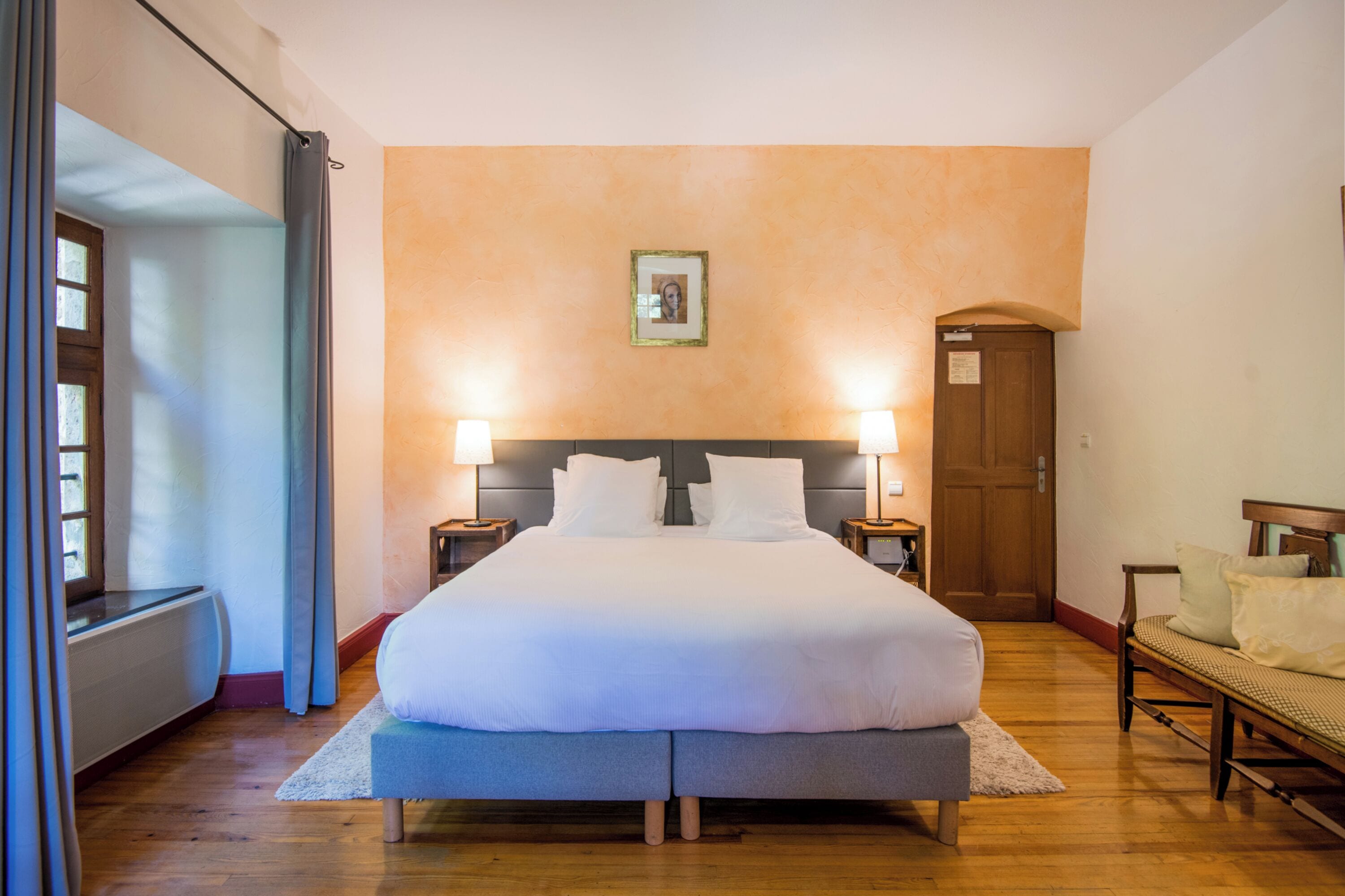 A hotel room with a large bed, two bedside lamps, a wooden bench, a door, and a framed picture on the wall.