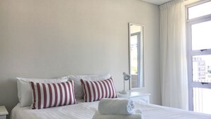 En pænt redt seng med stribede puder, en sengetavlelampe og et vindue med gardiner.