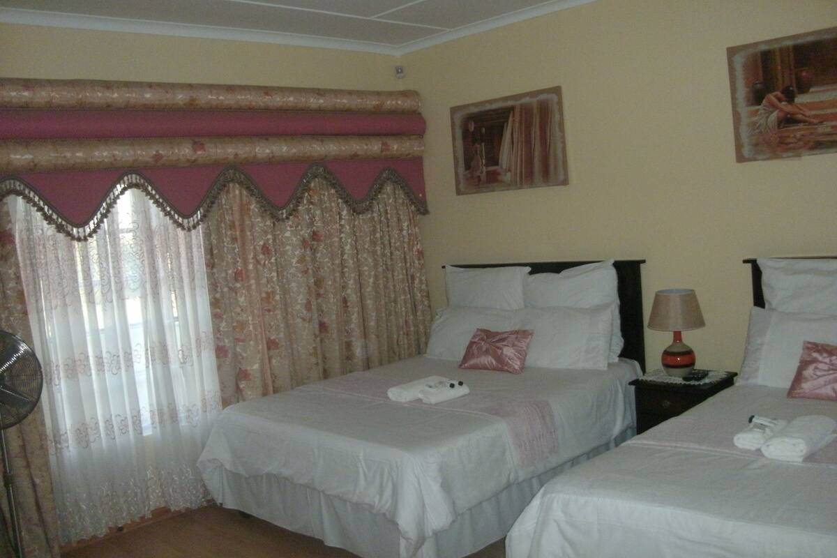 A hotel room with two beds, a bedside table with a lamp, a fan, and a painting on the wall.