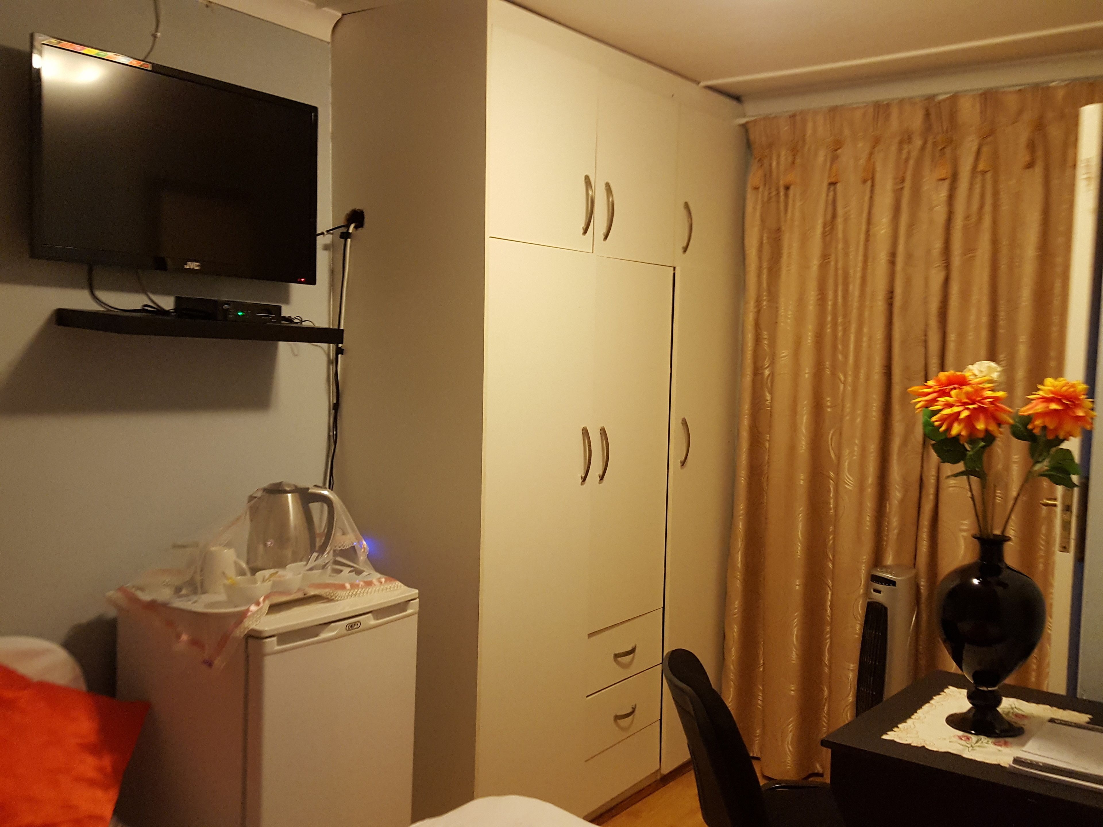 Habitación de hotel con televisor montado en la pared, minibar, escritorio con un florero y cama con almohada roja.