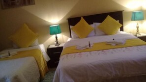 Quarto de hotel com duas camas, cada uma com colcha e travesseiros amarelos, uma mesa de cabeceira com abajur e uma luminária na parede.