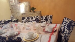 Uma sala de jantar com mesa posta para quatro pessoas, parede de pedra ao fundo, almofadas estampadas e um vaso de planta.