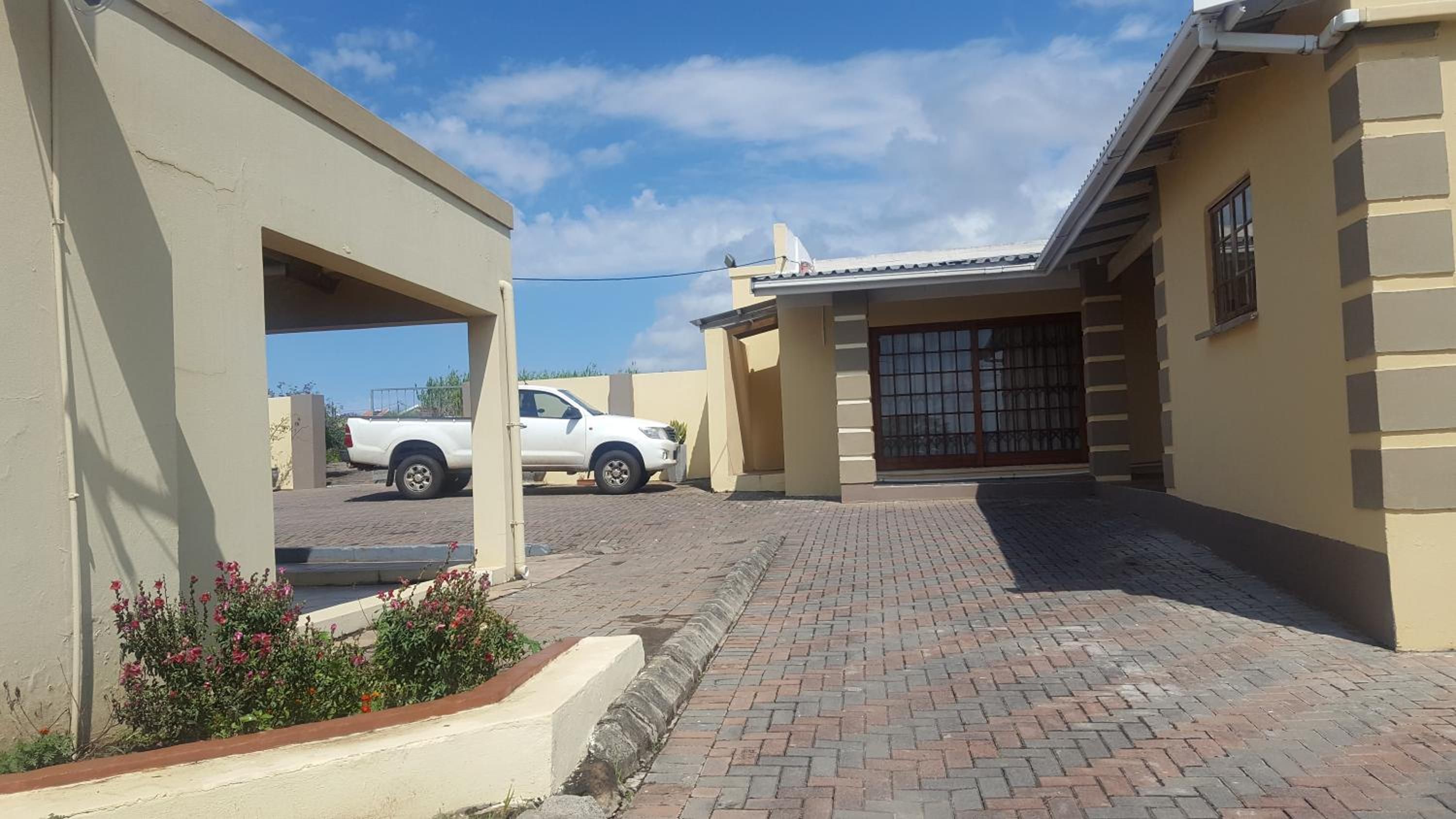 A paved driveway leading to a gated entrance, a white pickup truck parked in the background, and a small garden with pink flowers.